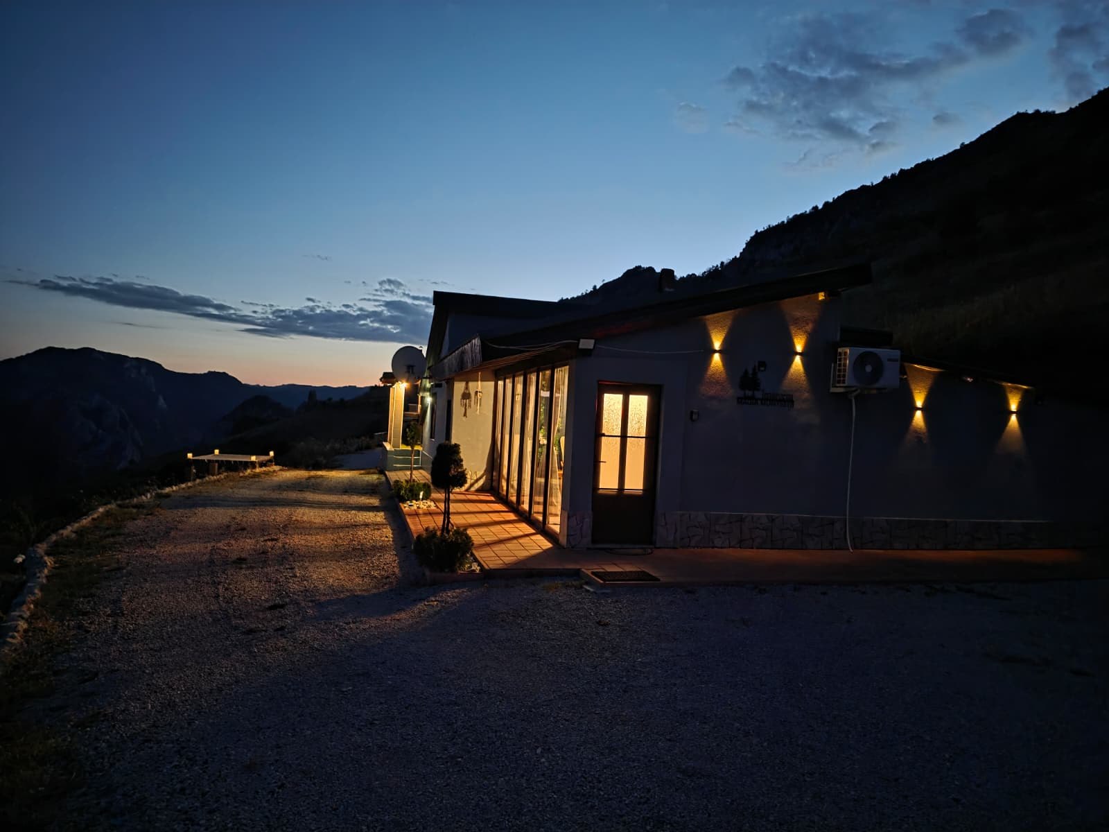 Casa iluminată la apus, cu peretele de sticlă spre terasă și cerul încă albastru peste creste